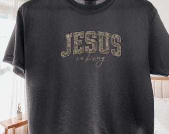 May include: A charcoal gray short-sleeve t-shirt featuring the text 'JESUS' in a block font with a camouflage pattern and the word 'is King' in a cursive script font centered on the chest. The shirt is displayed on a wooden hanger.