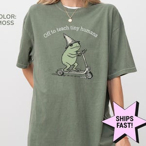 May include: Moss green t-shirt with the text "Off to teach tiny humans." The shirt features a cartoon frog wearing a wizard hat and riding a scooter. The frog is light green, and the scooter is white.