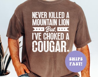 May include: Brown t-shirt with the text "NEVER KILLED A MOUNTAIN LION But, I'VE CHOKED A COUGAR." in white. The shirt has a crew neck and short sleeves. The text is in a bold, sans-serif font.