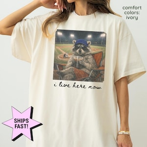 Può includere: T-shirt color avorio con una grafica di un procione con occhiali da sole e berretto da baseball, seduto su una sedia da stadio con una bevanda. Il testo "i live here now" è sotto l'immagine. Le parole "comfort colors: ivory" sono stampate in alto a destra.