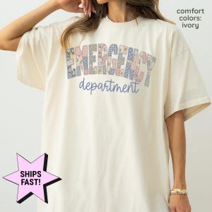 May include: An ivory short-sleeve t-shirt featuring the text EMERGENCY in a multi-colored patchwork floral and gingham pattern, with the word department written below in a simple blue script font. The shirt includes the text comfort colors: ivory.