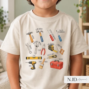 Puede incluir: Una camiseta beige de manga corta con un estampado gráfico de varias herramientas, incluyendo un martillo, un martillo de uña, un destornillador plano, un destornillador Phillips, una llave inglesa, una llave ajustable, alicates, una cinta métrica, un serrucho, un nivel, un taladro, una brocha, un cúter y una caja de herramientas roja.