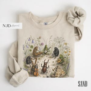 May include: A sand-coloured crewneck sweatshirt featuring a detailed graphic print of various insects playing musical instruments, including a xylophone, harp, banjo, and cello, surrounded by botanical illustrations of wildflowers and grasses.