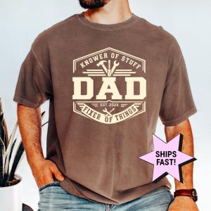 May include: A brown short-sleeve t-shirt featuring a cream-colored graphic with the text "KNOWER OF STUFF DAD FIXER OF THINGS EST. 2026" and icons of a hammer, screwdriver, and wrench arranged within a hexagonal border design.