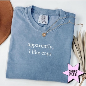 Puede incluir: Una camiseta azul Comfort Colors con el texto "apparently, i like cops" en blanco. Un collar dorado con un colgante descansa sobre la camiseta. La camiseta es talla S-P-CH.