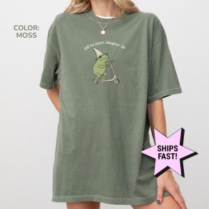May include: Moss green t-shirt with a frog riding a scooter graphic. The frog wears a wizard hat and the text "Off to start chapter 30" is above the frog. The shirt is a relaxed fit.