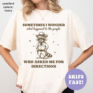 May include: Ivory-colored t-shirt featuring a raccoon wearing a cowboy hat and holding guns. The text reads "SOMETIMES I WONDER what happened to the people WHO ASKED ME FOR DIRECTIONS." The shirt is a comfort color.