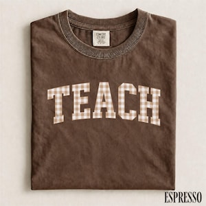 May include: A dark brown short-sleeve t-shirt with a crew neckline. The word TEACH is printed across the chest in a beige and white gingham pattern. The shirt features a Comfort Colors brand label on the inside of the collar at the neckline.