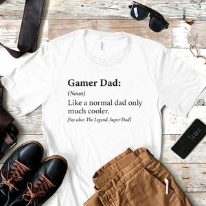 May include: White t-shirt with black text that reads "Gamer Dad: (Noun) Like a normal dad only much cooler. [See also: The Legend, Super Dad]"