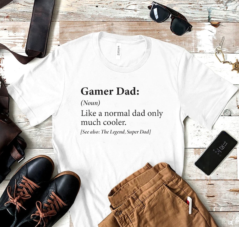 May include: White t-shirt with black text that reads "Gamer Dad: (Noun) Like a normal dad only much cooler. [See also: The Legend, Super Dad]"