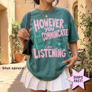 Puede incluir: Una camiseta de manga corta color azul abeto con el texto rosa "HOWEVER YOU COMMUNICATE i am LISTENING" junto a un gráfico de un walkie-talkie y un sobre con un corazón. La camiseta se combina con una falda plisada blanca y un bolso negro.