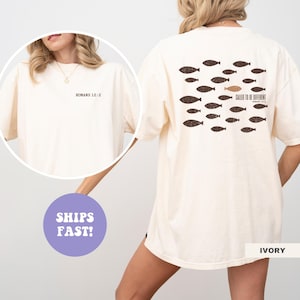 以下が含まれることがあります： アイボリーの半袖Tシャツ。左胸にROMANS 12:2と書かれた小さな黒いテキストがプリントされています。背面には、ダークブラウンの魚のシルエットのパターンと、1匹の金色の魚、そしてCALLED TO BE DIFFERENTというテキストがデザインされています。