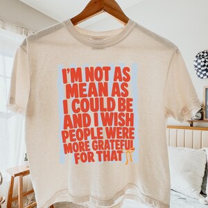 May include: An ivory boxy t-shirt featuring a light blue vertical striped background with red text that reads, I'M NOT AS MEAN AS I COULD BE AND I WISH PEOPLE WERE MORE GRATEFUL FOR THAT, accompanied by a small orange bow graphic.