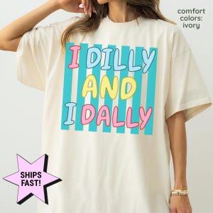 May include: An ivory short-sleeve t-shirt featuring a graphic design with vertical turquoise stripes. The text reads I DILLY AND I DALLY in a playful, rounded font with pink, light blue, and yellow lettering against the striped background.