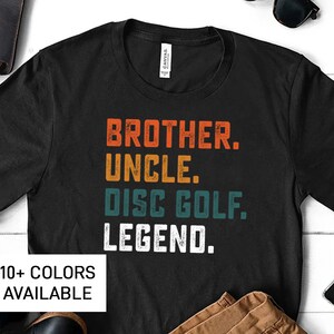Może przedstawiać: Czarny T-shirt z vintage'owym wzorem, na kt&oacute;rym widnieje napis "Brother. Uncle. Disc Golf. Legend." w kolorze białym, turkusowym i pomarańczowym. Tekst jest przetarty i ma wyblakły wygląd. Dostępnych jest ponad 10 kolor&oacute;w.