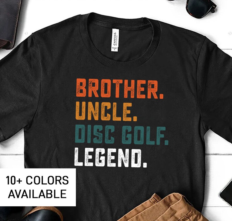 Może przedstawiać: Czarny T-shirt z vintage'owym wzorem, na kt&oacute;rym widnieje napis "Brother. Uncle. Disc Golf. Legend." w kolorze białym, turkusowym i pomarańczowym. Tekst jest przetarty i ma wyblakły wygląd. Dostępnych jest ponad 10 kolor&oacute;w.