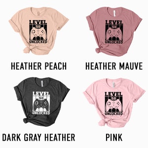 May include: Four t-shirts in different colors with a graphic of a video game controller and the text "Level 50 Unlocked". The colors are heather peach, heather mauve, dark gray heather, and pink.