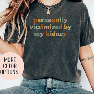 Peut inclure: Un t-shirt gris foncé avec le texte "personally victimized by my kidney" en lettres colorées. Le texte est dans un style de police rétro.
