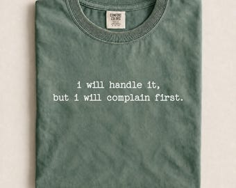 May include: A moss green crew neck t-shirt with a vintage-washed finish. The center of the chest features white typewriter-style text that reads: i will handle it, but i will complain first. The inner neck label displays the Comfort Colors brand logo.