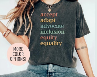 May include: A dark gray t-shirt with the words "accept", "adapt", "advocate", "inclusion", "equity", and "equality" printed in colorful letters. The words are stacked vertically on the shirt.
