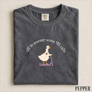 May include: A charcoal grey crew neck t-shirt featuring a graphic of a white goose wearing a headset and riding a purple scooter. The text above the goose reads, "off to answer some 911 calls" in white lettering. The shirt has a Comfort Colors label.