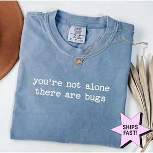 May include: A blue short-sleeved t-shirt with a crew neckline and a white printed graphic that reads, you're not alone there are bugs. The shirt features a Comfort Colors brand label at the inner neck and is paired with a gold-toned pendant necklace.