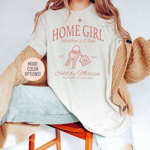 May include: Cream-colored t-shirt with the text "HOME GIRL Realtor's Club" in a coral color. The design includes keys and a house graphic, with the text "Sold by Melissa" and "Your Company Name Here". The shirt also has "EST. 2024" printed on it.
