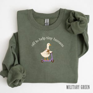 May include: A military green crewneck sweatshirt featuring a graphic of a white goose riding a purple scooter while holding a brown teddy bear. The text above the goose reads, off to help tiny humans, in a white, curved, sans-serif font.