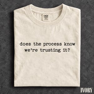 Puede incluir: Una camiseta de cuello redondo color marfil con el texto impreso en negro "does the process know we're trusting it?" en el pecho. La camiseta tiene una etiqueta de marca visible en el interior del cuello y un corte estándar de manga corta.