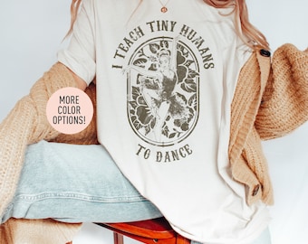 May include: Cream-colored t-shirt with the text "I TEACH TINY HUMANS TO DANCE" in a circular design. The design features a ballerina and floral elements. The shirt is paired with light-wash jeans and a tan cardigan. A button on the cardigan is visible.