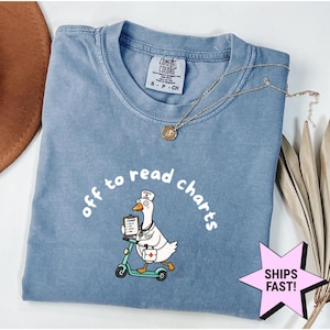May include: A blue short-sleeve t-shirt featuring a graphic of a goose dressed as a medical professional riding a scooter. The goose wears a nurse cap and stethoscope, holding a clipboard and medical bag. The text reads, off to read charts.