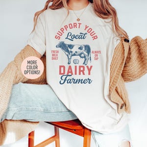 May include: A white t-shirt with a vintage-style graphic design that says "Support Your Local Dairy Farmer". The graphic features a black and white cow, a barrel, and the text "Fresh Milk Daily" and "Always On Tap".
