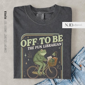 May include: A charcoal gray t-shirt featuring a graphic of a green frog wearing a sweater and riding a bicycle. The frog carries a basket of books and a lamp. The text reads OFF TO BE THE FUN LIBRARIAN. The shirt has a crew neck and short sleeves.