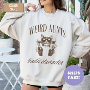 May include: Sand-coloured sweatshirt with the text "WEIRD AUNTS build character." The graphic features a cat wearing sunglasses and holding wine glasses. The sweatshirt is a crew neck style.