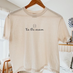 May include: An ivory boxy t-shirt featuring a black graphic of a baseball and the text 'tis the season centered on the chest. The shirt has a crew neckline, short sleeves, and a straight hemline, hanging from a wooden hanger against a neutral background.