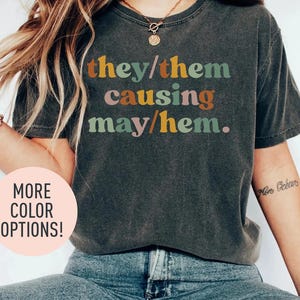 May include: A dark grey t-shirt with the text "they/them causing may/hem." printed in rainbow colours.