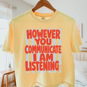 Op de afbeelding: Een lichtgeel T-shirt met korte mouwen, voorzien van een lichtblauwe rechthoekige achtergrond met verticale strepen en de rode tekst "HOWEVER YOU COMMUNICATE I AM LISTENING" met een klein oranje strikicoon aan het einde.