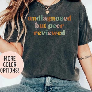 Può includere: Una maglietta grigia scura con il testo "undisagnosed but peer reviewed" in lettere colorate. Il testo è in uno stile di font retrò.