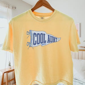 May include: A pale yellow short-sleeve t-shirt featuring a blue and white gingham pennant graphic on the chest. The pennant contains the text COOL AUNT in bold blue letters, accented with small blue ribbons on the left side and a star on the right.