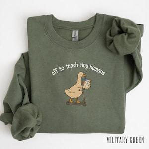 Peut inclure: Un sweat-shirt à col rond vert militaire orné d'un motif représentant une oie brun clair sur une trottinette, tenant une pile de livres. Le texte au-dessus de l'oie indique "off to teach tiny humans" dans une police blanche arrondie.