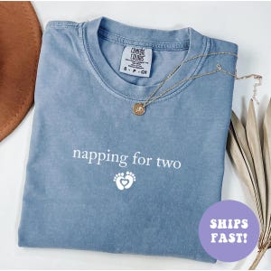 May include: A blue Comfort Colors t-shirt with the text "napping for two" and a footprint graphic. A gold necklace with a pendant rests on the shirt. The shirt is folded and has a tag that says "S - P - CH".