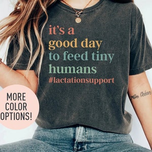 Puede incluir: Una camiseta gris oscuro con el texto "it's a good day to feed tiny humans #lactationsupport" impreso en colores arcoíris.