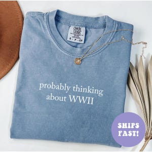 May include: A blue Comfort Colors t-shirt with the text "probably thinking about WWII" in white. A gold necklace with a pendant rests on the shirt. The shirt is folded and has a small tag.