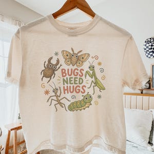 May include: An ivory boxy t-shirt featuring a graphic print of a moth, beetle, praying mantis, and caterpillar surrounding the text BUGS NEED HUGS in red and green. The shirt has a crew neckline and short sleeves, displayed on a wooden hanger.