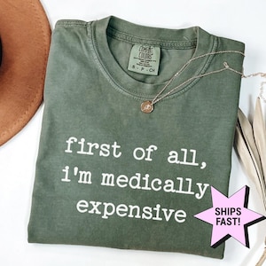 May include: Olive green Comfort Colors t-shirt with the text "first of all, I'm medically expensive" in white. A gold necklace with a pendant is draped on the shirt. A pink starburst graphic with "SHIPS FAST!" is in the lower right corner.