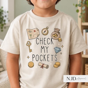 May include: A beige t-shirt featuring a graphic print with the text "CHECK MY POCKETS" surrounded by illustrations of a treasure map, a compass, a bag of gold coins, a key, a diamond, a scroll, and additional loose gold coins.