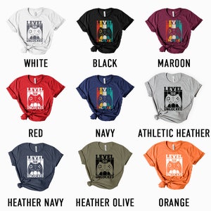 May include: A mockup of a t-shirt design with the text "Level 50 Unlocked" and a retro-style video game controller graphic. The shirt is available in 10 different colors: white, black, maroon, red, navy, athletic heather, heather navy, heather olive, and orange.