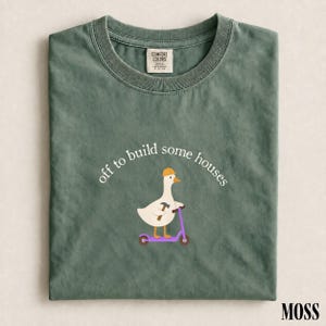 Può includere: Una t-shirt girocollo color verde muschio con la grafica di un'anatra bianca che indossa un casco da cantiere giallo, tiene in mano un martello e cavalca un monopattino viola. Il testo sopra l'anatra recita, off to build some houses.