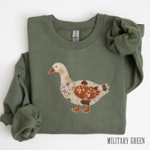 Peut inclure: Un sweat-shirt à col rond vert olive orné d'un motif central représentant une oie. L'oie est composée de divers empiècements en tissu à motifs dans des tons crème, orange et marron, incluant des motifs floraux et solaires. Le texte indique Military Green.