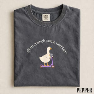 May include: A charcoal grey t-shirt featuring a graphic of a white goose riding a purple scooter while holding a calculator and a pencil. The text above the goose reads, "off to crunch some numbers." The shirt has a Comfort Colors brand label.
